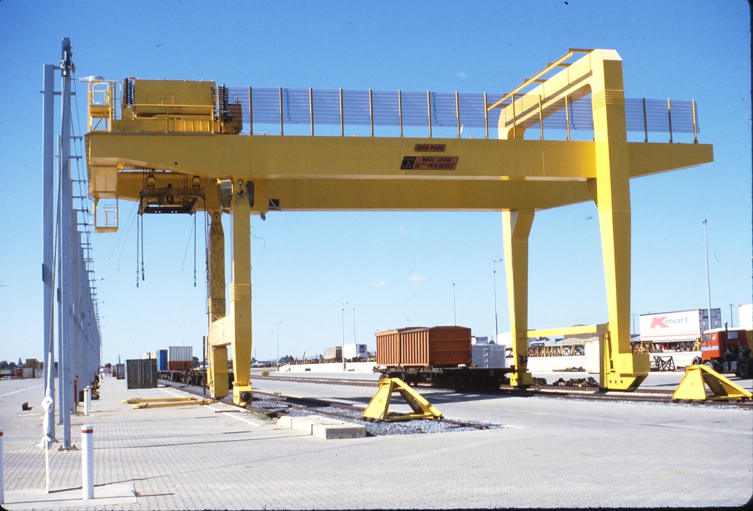 En stor gul portalkran vid vad som verkar vara en intermodal godsterminal eller containergård. Bilden visar infrastruktur som används för hantering av fraktcontainrar i ett logistiknav, med synliga järnvägsspår. Detta representerar en kritisk nod i urbana logistiknätverk där varor överförs mellan olika transportsätt före slutdistribution i stadsområden.