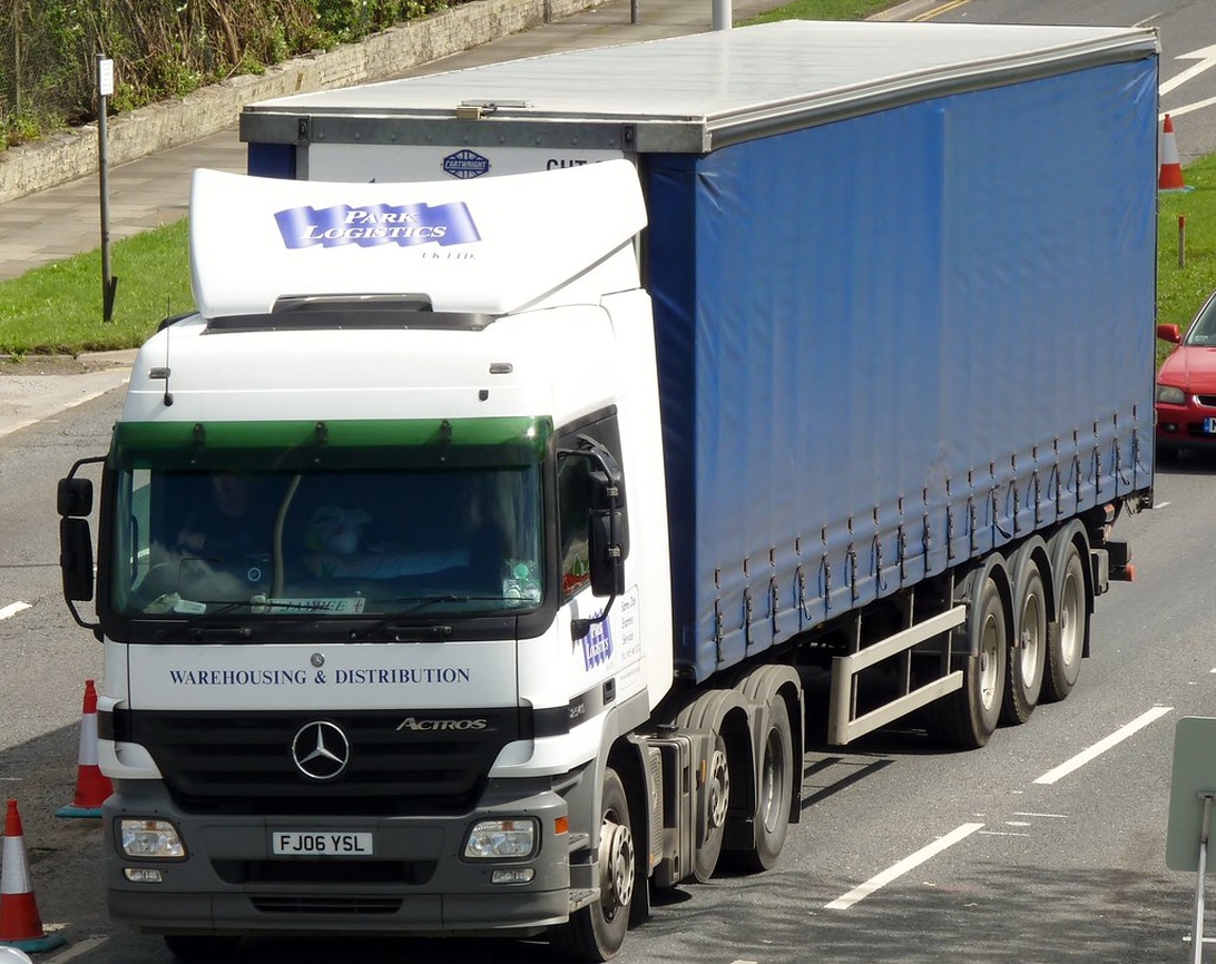 En Mercedes Actros lastbil med 'Park Logistics' märkning parkerad på en stadsgata. Det stora blå kapellsläpet är tydligt märkt med texten 'Warehousing & Distribution', vilket representerar en nyckelkomponent i urban logistik - transport av varor inom stadsmiljöer. Fordonet är placerat på vad som verkar vara en leveransrutt i en urban miljö.
